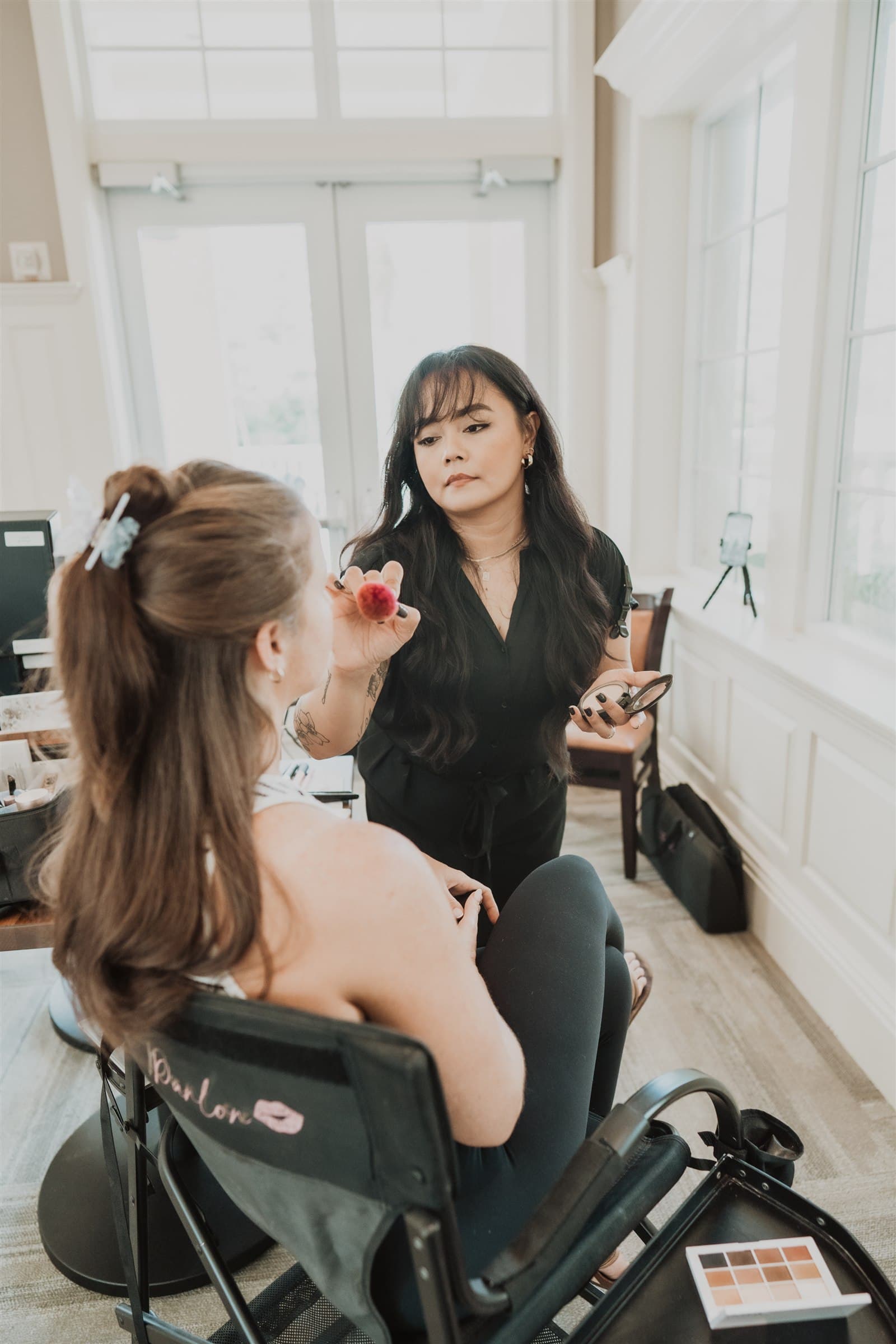 Maureen applying bridal makeup beside a chair embroidered with The Parlor