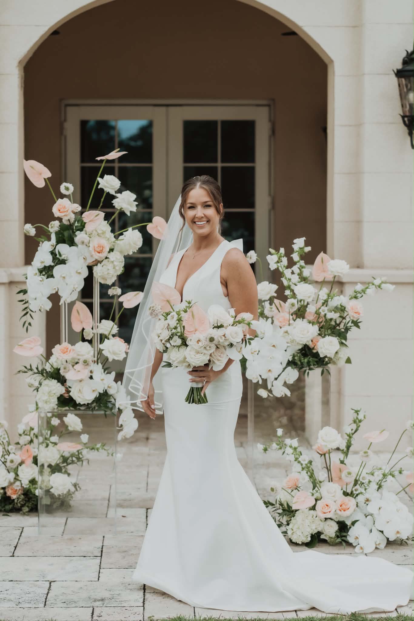 Bride with a soft updo and glowing makeup holding her bouquet in front of a floral ceremony arch